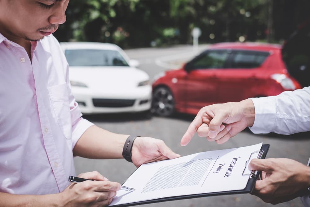 After a traffic accident, an insurance agent inspects a damaged car, while the customer signs the report claim form.