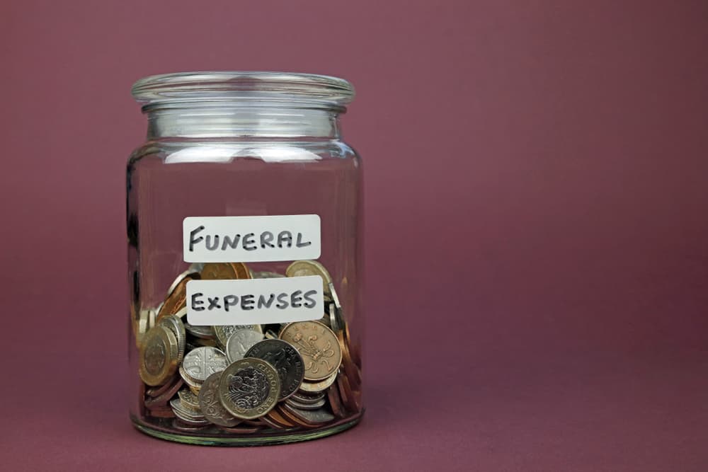 Glass jar labeled 'Funeral Expenses' filled with money, symbolizing funds set aside for funeral costs.