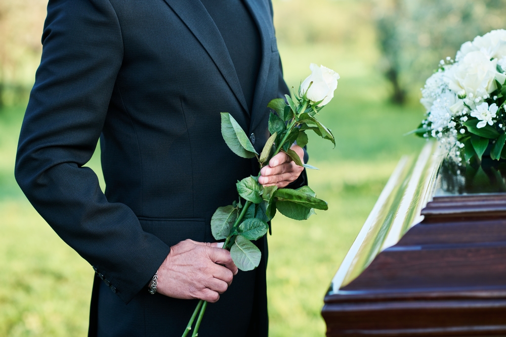 A person in a black suit holding a white rose, standing solemnly by a casket adorned with flowers, symbolizing a wrongful death and the grieving process.