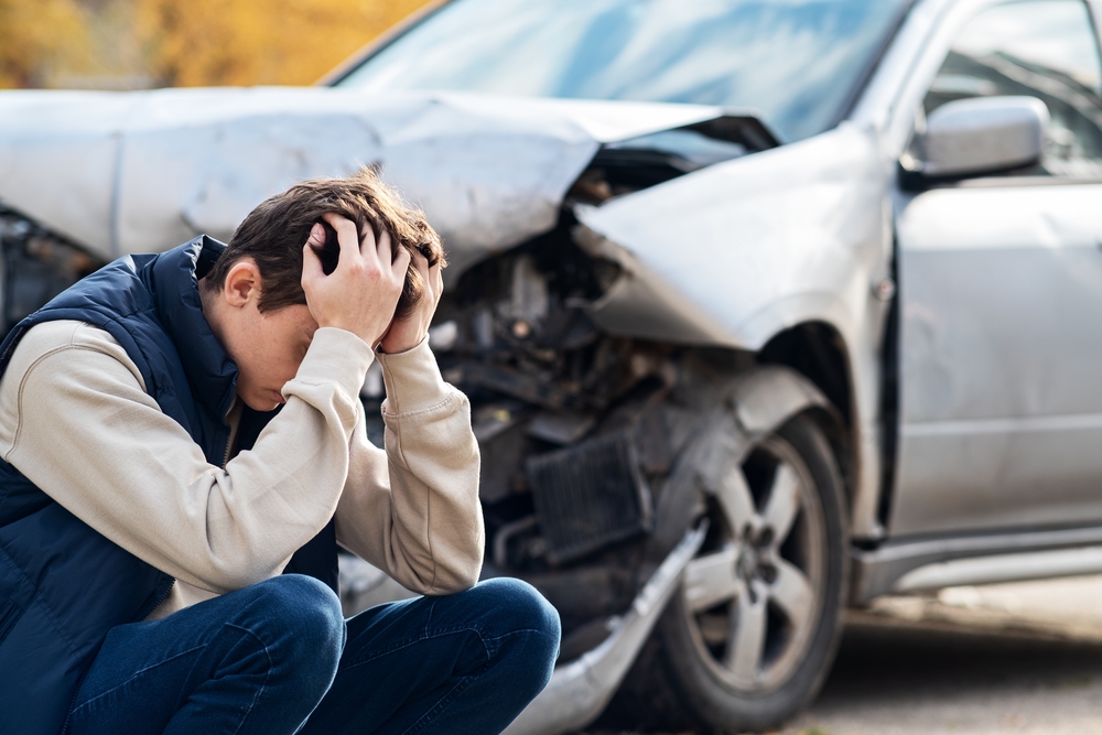 A frustrated man stands beside a severely damaged car, hands on his head, realizing the extent of the damage is irreparable