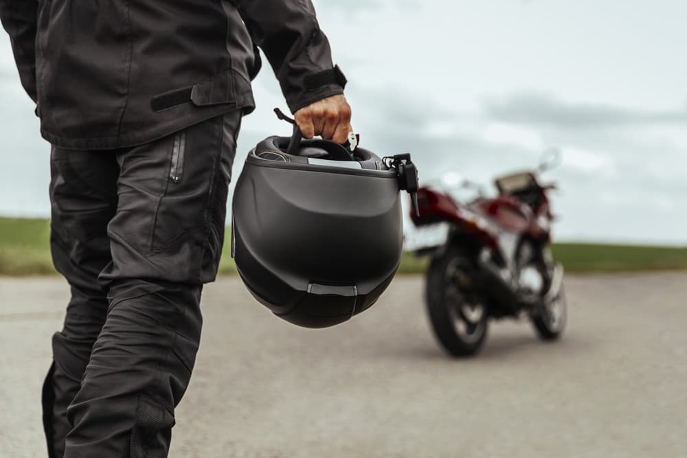 Motorcyclist walking along a road while carrying a motorcycle helmet, with a motorcycle parked in the background