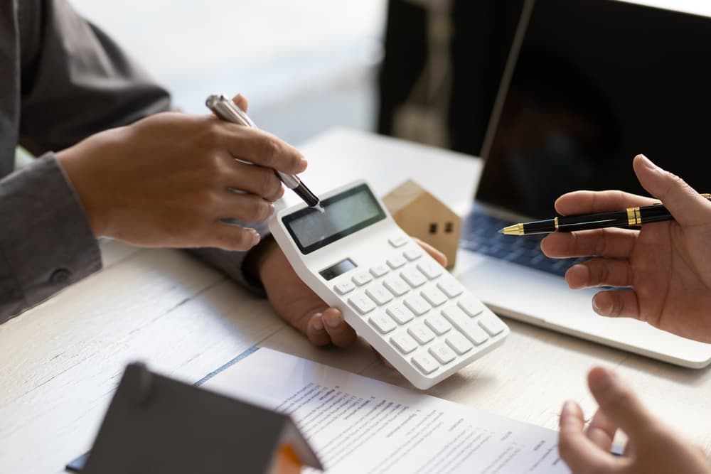 Two individuals reviewing financial documents and using a calculator during a meeting, with a laptop and small house model visible on the table