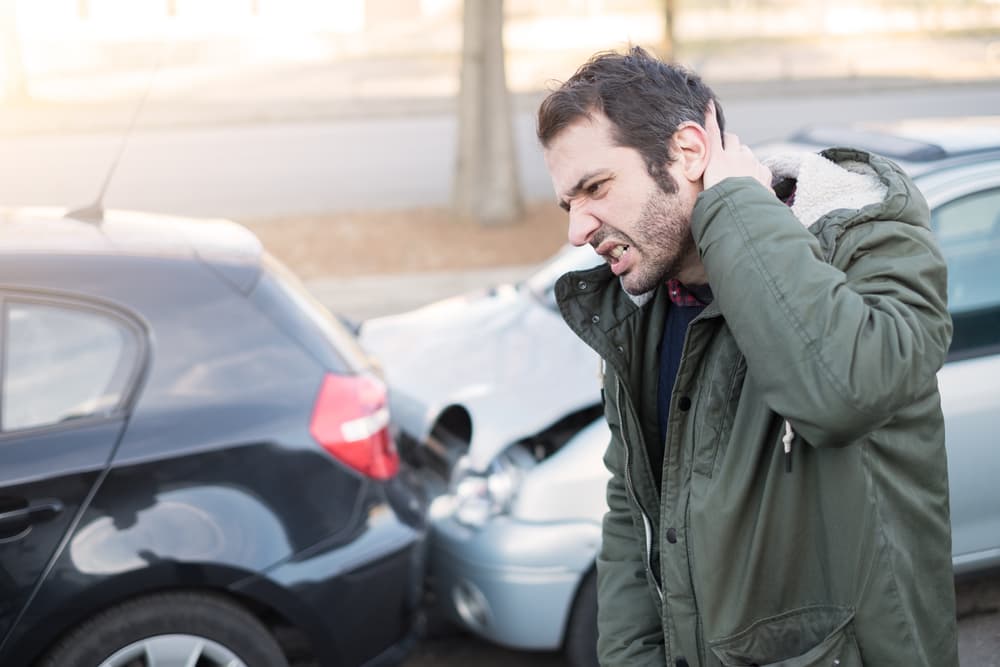 Man holding his neck in pain while standing beside two damaged vehicles after a rear-end car accident