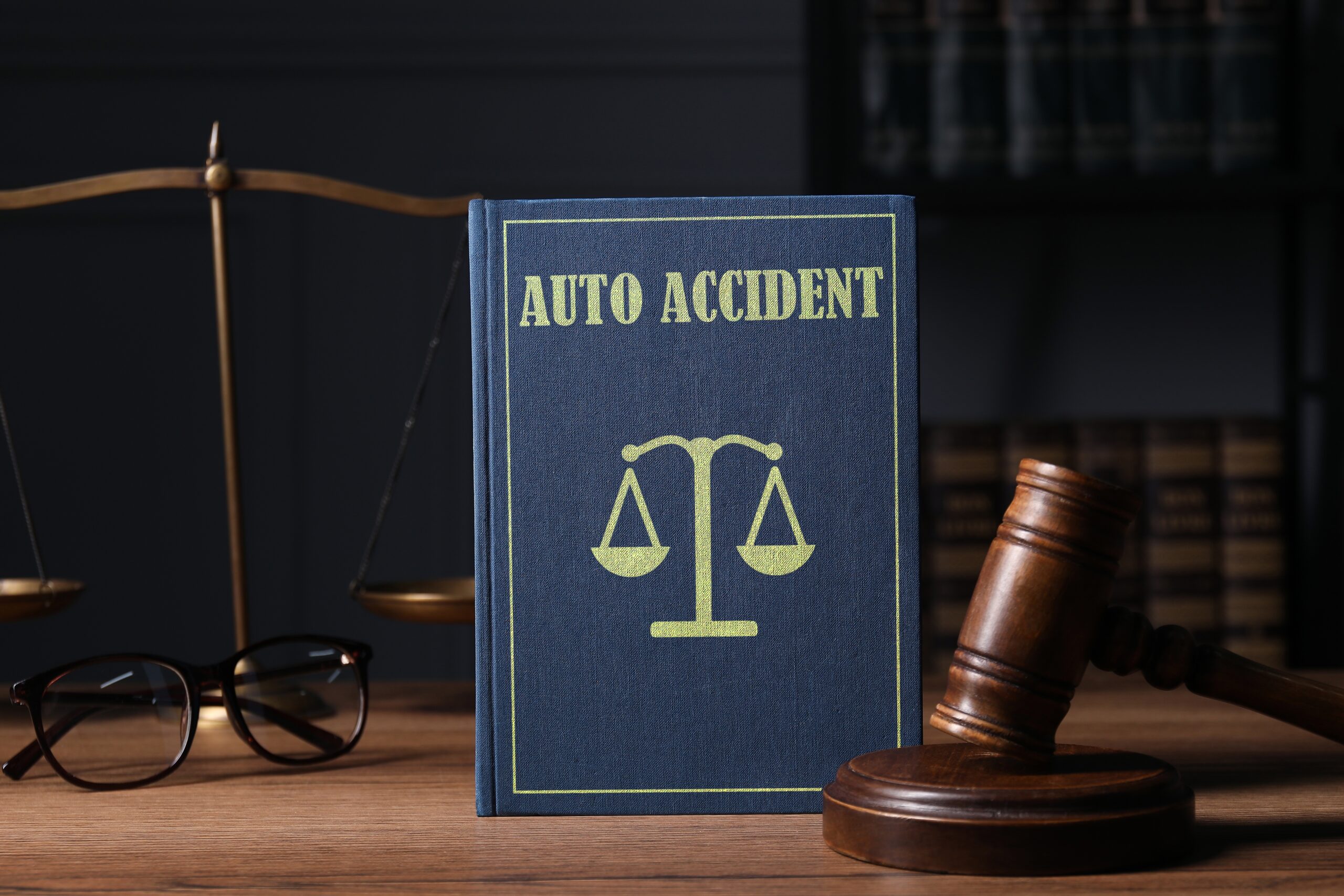 Book labeled &lsquo;Auto Accident&rsquo; displayed on a desk beside a judge&rsquo;s gavel, scales of justice, and eyeglasses in a law office setting